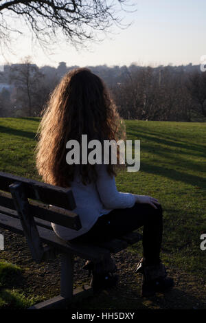 Nachdenkliche junge Frau sitzt allein auf einer Bank, Blick in die Ferne, in einer ruhigen Lage mit einem Himmel und Bäume. Stockfoto