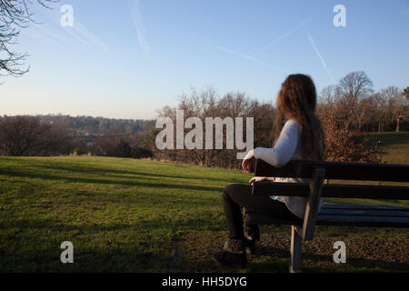 Nachdenkliche junge Frau sitzt allein auf einer Bank, Blick in die Ferne, in einer ruhigen Lage mit einem hellen Himmel und Bäume. Stockfoto