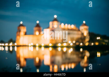 Mir, Weißrussland. Verschwommene Boke Bokeh Fokus von Mir Burganlage In Nachtbeleuchtung und Reflexionen auf See Wasseroberfläche In warmen gelben Farben. Stockfoto