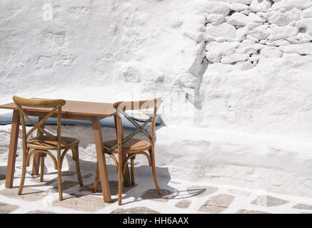 Ein leerer Außentisch mit zwei Stühlen steht in der griechischen Sonne auf einer weiß getünchten Terrasse. Stockfoto