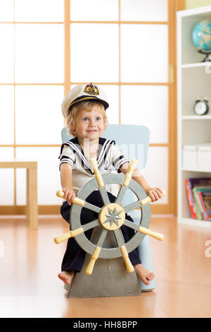 Kind als Kapitän oder Seemann verkleidet spielt auf Stuhl als auf Schiff in seinem Zimmer. Kind junge dreht das Holzlenkrad. Stockfoto