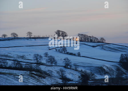 Sonnenuntergang fängt an und gibt den Fenstern eines Bauernhauses in einer winterlichen schneebedeckten Landschaft in Yorkshire ein goldenes Leuchten Stockfoto