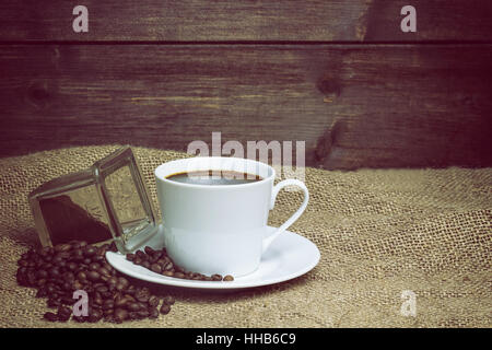 Weiße Tasse Kaffee auf der Sackleinen steht. Um ihn herum verstreut sind, Kaffeebohnen und gemahlenen Kaffee Glas Stockfoto