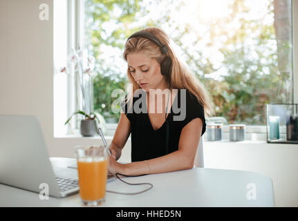 Ernste junge Frau Notizen während des Sitzens vor der Laptop-Computer am Küchentisch. Stockfoto
