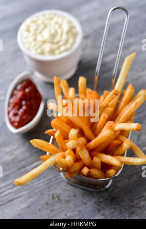 einige appetitlich Pommes Frites mit Mayonnaise und Ketchup auf einem grauen rustikalen Holztisch in einen Metallkorb und einige Schalen serviert Stockfoto