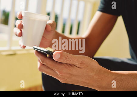 Closeup von einem kaukasischen Jüngling mit einer Tasse Kaffee in der Hand und sein Smartphone in die andere, auf einer Terrasse oder einem Balkon Stockfoto