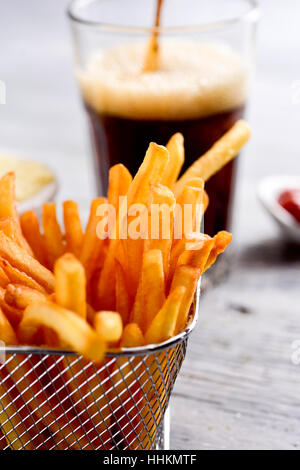 Nahaufnahme von einigen leckeren Pommes frites serviert in einem Metallkorb, ein Glas mit Soda und eine Schüssel mit Ketchup auf einem grauen rustikalen Holztisch Stockfoto