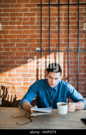 Junger Mann, Zeitung lesen, Kaffee zu geniessen, an einem Holztisch im Hipster-Café sitzen und blickte, Telefon und Gläser auf Tisch Stockfoto