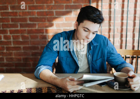 Junger Mann Lesung Papier und genießen Kaffee sitzen an einem Holztisch im Hipster-Café, Mobilgerät auf Tisch Stockfoto