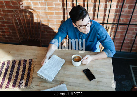 Junger Mann Lesung Papier und genießen Kaffee an einem Holztisch im Hipster-Café sitzen und Blick auf die Seite, Telefon auf Tisch Stockfoto