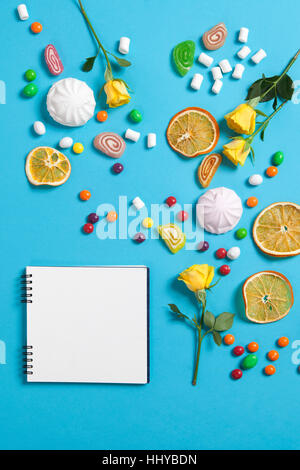 Marshmallows, Süßigkeiten, Gummibärchen, Süßigkeiten, trockenen Becher Orangen und gelben Rosen fallen auf einen Notizblock auf blauem Grund. Werbeflächen Stockfoto