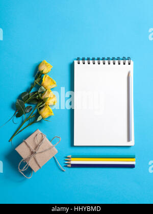 Quadratische Notizblock auf Federn mit weißen Kraftpapier, Bleistift und eine gelbe Rose lag auf einem blauen Hintergrund. Exemplar Stockfoto