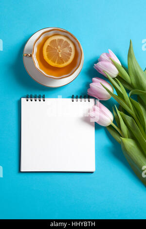 Quadratische Notizblock auf Federn mit weißen Kraftpapier, rosa Tulpen und Tasse Tee sind auf blauem Grund. Exemplar Stockfoto