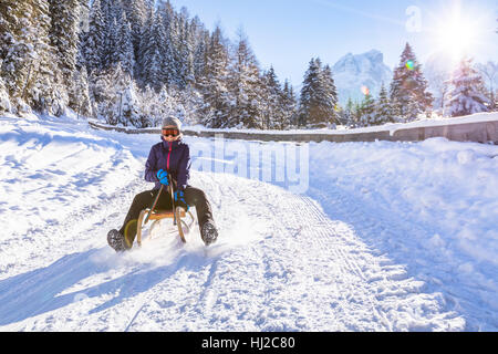 Fröhliches Mädchen reitet einen Schlitten bergab auf einer Schnee bedeckten Schlitten Trail in einer weißen sonnigen Winter Berglandschaft