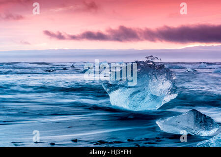 Eisberg entlang der Ufer der Jökulsárlón Gletscherlagune während einer pulsierenden roten Sunrise liegt bewegungslos, wie es von einer kalten Ozeanwasser umrahmt ist. Stockfoto