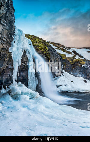 Ein kalten verschneiter Wasserfall im Hochland von Island umrahmt von Pastell Himmel und zerklüftete Gelände bietet landschaftlich Inbegriff der gefrorenen Wildnis Stockfoto