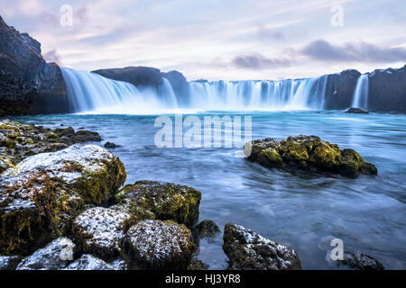 Godafoss Wasserfall bei Sonnenaufgang zeigt das Wasser in Strömen über die Kante und kicking eine neblige Wolke über dem Wasser mit Schnee bedeckt die Klippen. Stockfoto