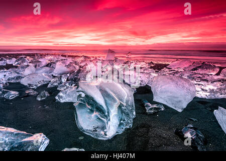 Eisberg entlang der Ufer der Jökulsárlón Gletscherlagune während einer pulsierenden roten Sunrise liegt bewegungslos, wie es von einer kalten Ozeanwasser umrahmt ist. Stockfoto