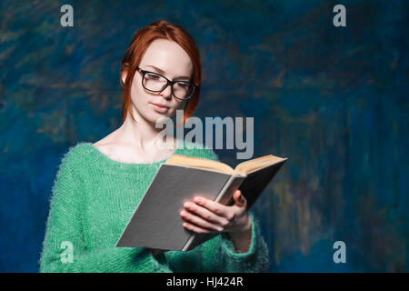 schöne rothaarige Lehrerin im grünen Pullover und Brille Buch über Magie dunkelblauen Hintergrund Stockfoto