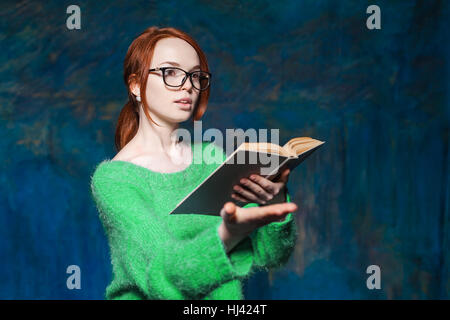 schöne rothaarige Lehrerin im grünen Pullover und Brille lesen Buch und Gesten über magische dunkelblauen Hintergrund Stockfoto