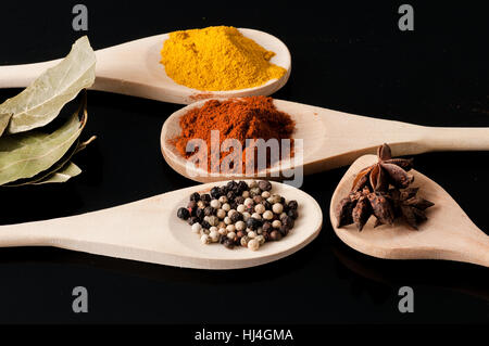 Hölzerne Löffel gefüllt mit Gewürzen auf schwarzem Hintergrund Stockfoto