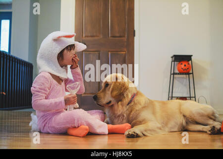 Mädchen im Bunny-Kostüm auf Boden essen Halloween Süßigkeiten mit Golden Retriever Hund sitzend Stockfoto