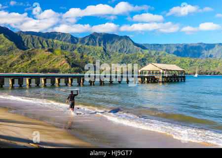 Surfer trägt Surfbrett im Hanalei Beach in der Nähe von Hanalei Pier auf Kauai Stockfoto