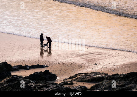 Zwei Menschen und ihrem Hund in der Silhouette zu sehen, wie die Sonne über Fistral Beach in Newquay, Cornwall. Stockfoto