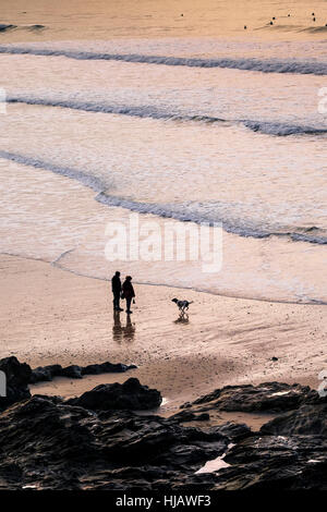 Zwei Menschen und ihrem Hund in der Silhouette zu sehen, wie die Sonne über Fistral Beach in Newquay, Cornwall. Stockfoto