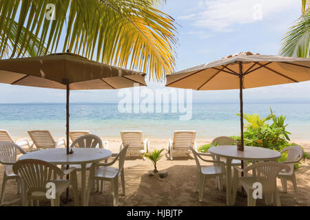 Restauranttische, Liegestuhl und Sonnenschirm am Strand unter Palmen am Strand verlässt Stockfoto