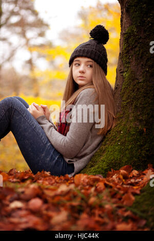 Hübsches junges Mädchen sitzt neben einem Baum im Herbst mit einem Teppich, wenn Blätter auf dem Boden Stockfoto