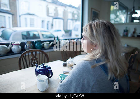 Junge Frau sitzt im Café, Tasse Tee und Teekanne auf Tisch, Blick aus Fenster Stockfoto