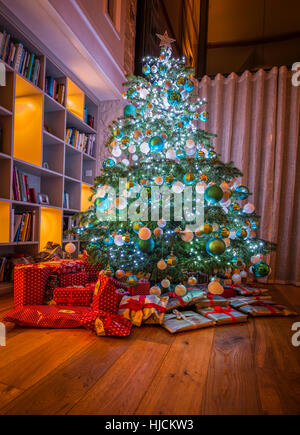 Großer Weihnachtsbaum mit verpackte Geschenke/Geschenke in Haus in England UK. Baum hat Weihnachtsbeleuchtung und Kugeln. Bücherregale in B/G Stockfoto