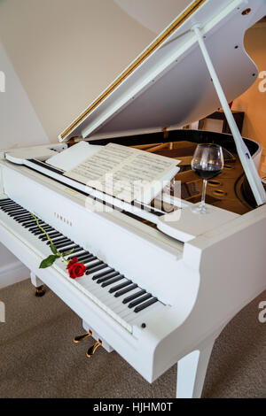 Weiße Flügel mit einem roten rose auf der Tastatur ein Glas Rotwein und ein Notenblatt zum feiern einen besonderen Anlass Stockfoto