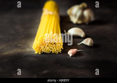 Bündel von ungekochten Spaghetti eng mit Bindfaden auf schwarzen zerkratzt konkrete Hintergrund mit verstreuten Knoblauchzehen, schließen sich Stockfoto