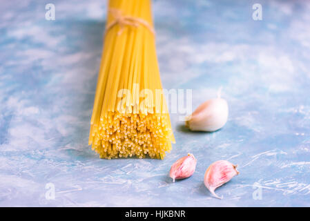 Bündel von ungekochten Spaghetti eng mit Bindfaden auf blauen und weißen konkrete Hintergrund mit verstreuten rosa Knoblauchzehen, Nahaufnahme Stockfoto