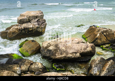 Schöne Szene mit Steinen mit grünem Moos an der Küste, Meerwasser, das zwischen ihnen, einige kleine Wellen und Leute schwimmen im Hintergrund. Stockfoto