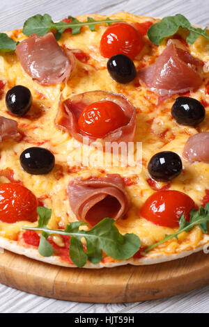 Italienische Pizza mit Schinken, Tomaten, schwarzen Oliven und Rucola-close-up auf einem Holztisch. vertikale Stockfoto