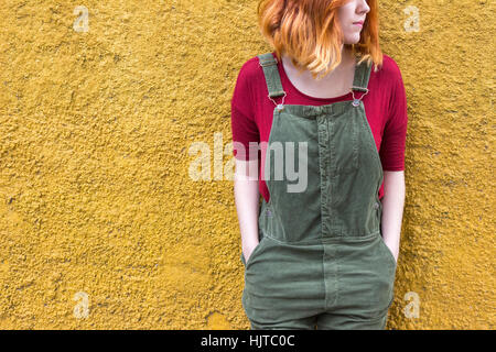 Junge Frau mit roten Kurzhaar und tragen grüne Overalls mit einem roten Hemd Lächeln stehend gegen gelb Stockfoto