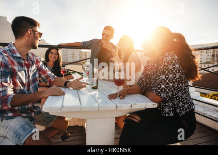 Gruppe von jungen Leuten an einem Tisch mit Getränken. Junge Männer und Frauen, die auf dem Dach Abend feiern. Stockfoto
