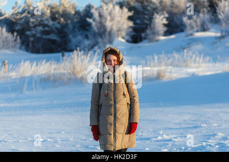 Junge fröhliche Frau zu Fuß in der kalten sonnigen Wintertag auf dem Hintergrund der Bäume und Schnee. Mädchen in warmen Beige Jacke rote Handschuhe Stockfoto