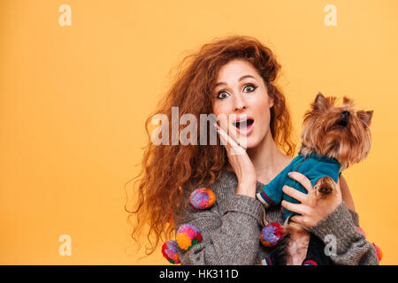 Junge rote Kopf Frau überrascht halten Sie ihren Hund auf orangem Hintergrund isoliert Stockfoto