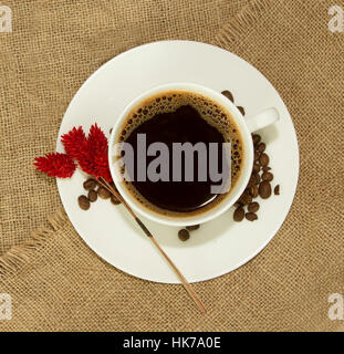 Weiße Tasse mit Kaffeebohnen auf Untertasse mit trockene rote Blume auf Leinentuch Stockfoto