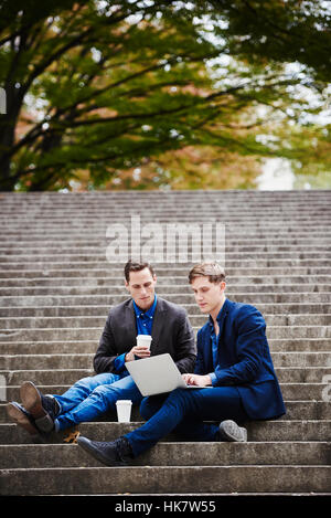 Zwei junge Männer sitzen auf einer Treppe im freien Blick auf einen Laptop zusammen. Stockfoto