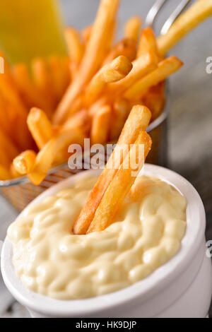 Nahaufnahme einer weißen Keramik Schüssel Mayonnaise mit einigen leckeren Pommes frites serviert in einem Metallkorb auf einem grauen rustikalen Holztisch Stockfoto