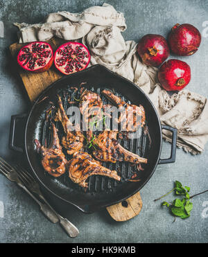 Barbecue-Dinner. Gegrillte Lammkoteletts mit Zwiebeln und Rosmarin in schwarzem Gusseisen Pfanne Fleisch serviert mit frischen Granatäpfeln auf Holzbrett über graue concr Stockfoto