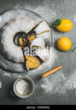Hausgemachte glutenfreie Zitronen Gugelhupf mit Puderzucker serviert mit frisch gepflückten Zitronen auf grauem Beton Hintergrund, Draufsicht, vertikale compositio Stockfoto
