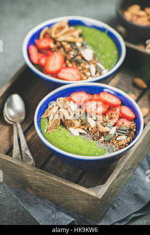 Gesundes Frühstück. Grüner Smoothie Schalen mit frischen Erdbeeren, Müsli, Chia und Kürbis Samen, Trockenfrüchten und Nüssen in Holztablett über graue Bereitung Stockfoto