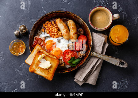 Englisches Frühstück in Pfanne mit Spiegelei, Würstchen, Speck, Bohnen, Toast und Kaffee auf dunklen Stein Hintergrund Stockfoto