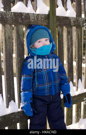 entzückende Kleinkind Junge stand gegen Zaun tragen Winter Jacke Mütze Schal spielen im Schnee Stockfoto